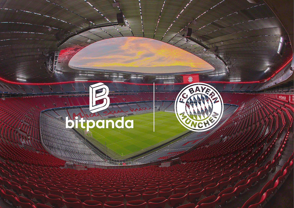 Interior of a large, empty stadium with red seats, featuring Bitpanda and FC Bayern München logos on the field, under a sky-lit roof.