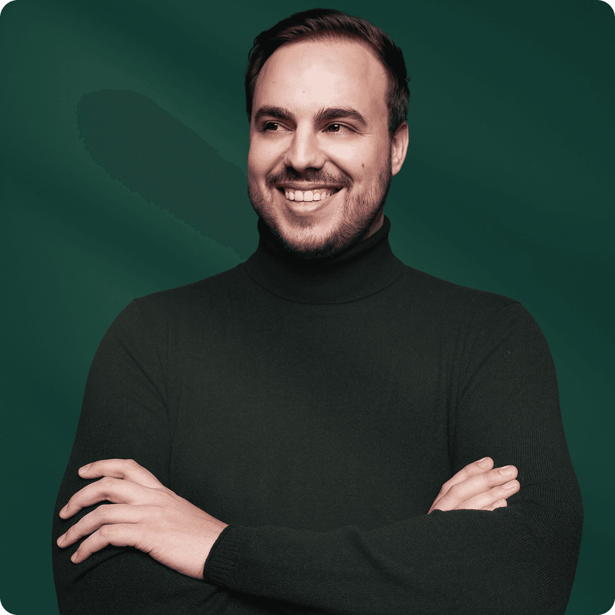 Smiling man in a dark turtleneck with arms crossed, standing against a dark green background.