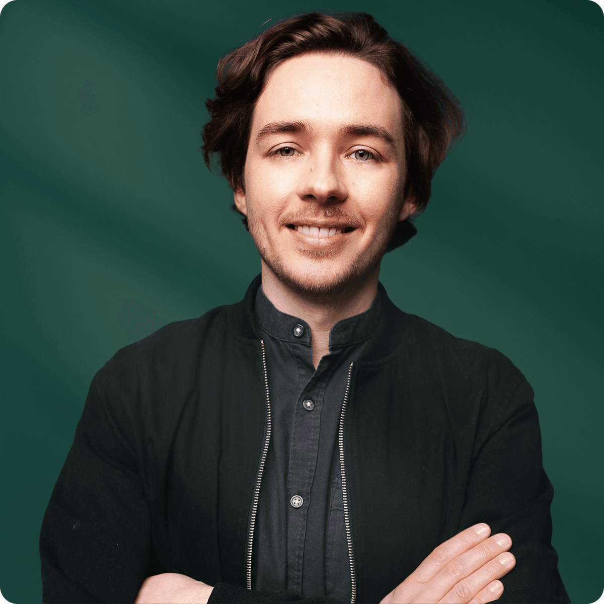 Smiling person with brown hair, wearing a dark jacket and shirt, stands with arms crossed against a green background.