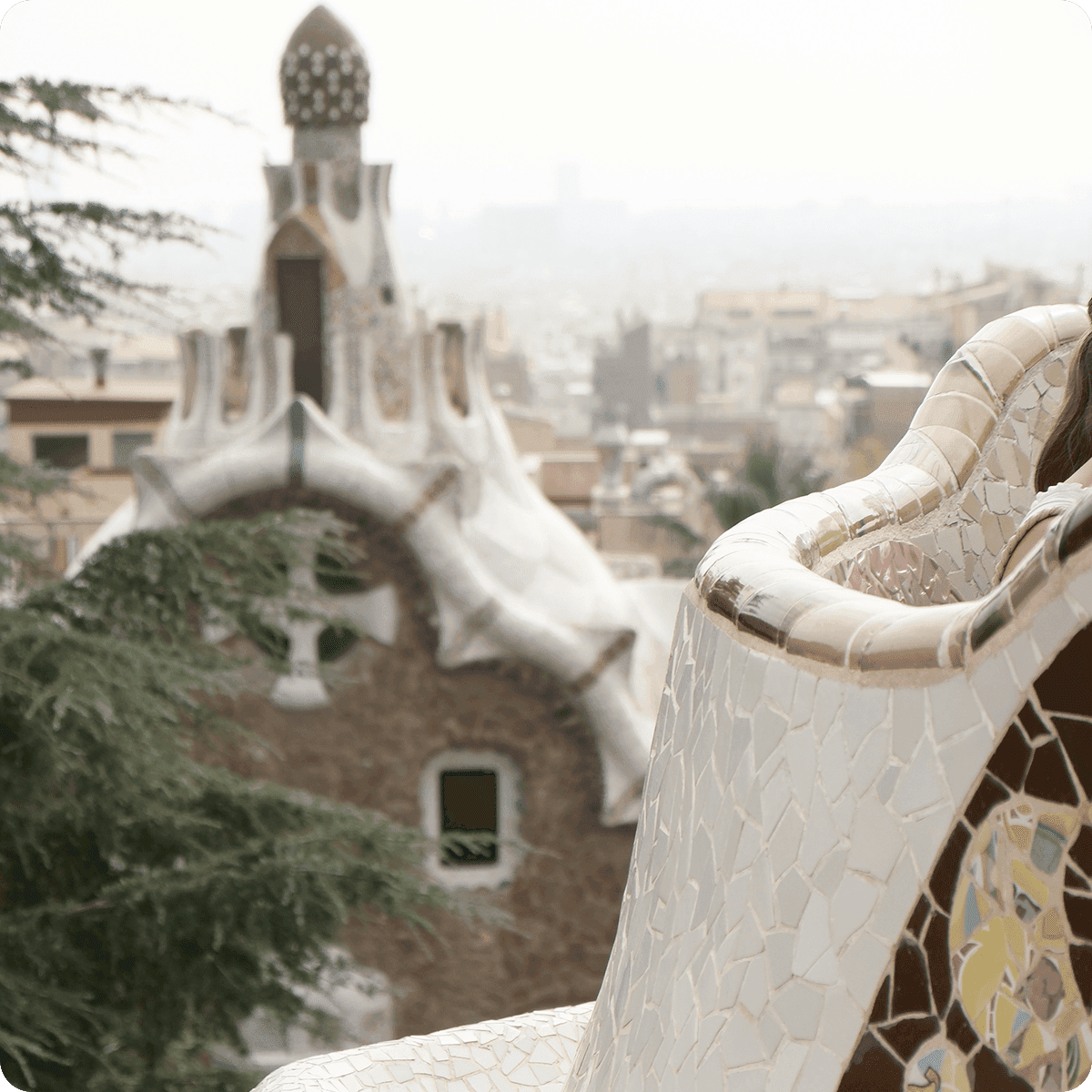 Mosaic bench with colorful tiles in Park Güell, Barcelona, overlooking a cityscape and a unique, ornate building in the background.