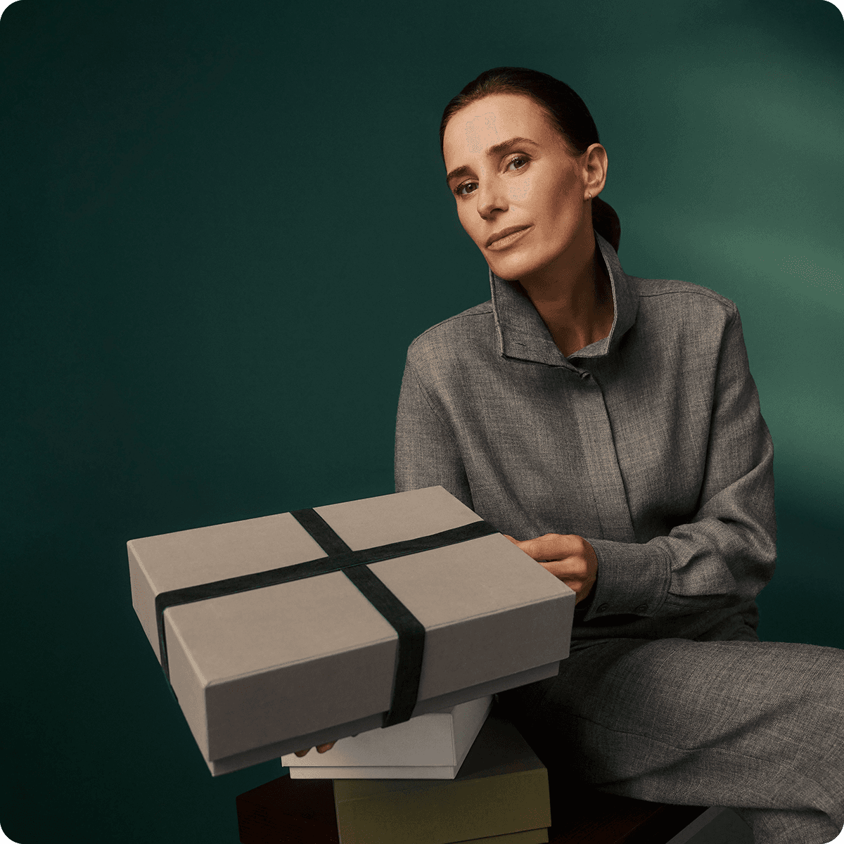 Woman in a gray outfit holding a gift box with a dark green ribbon, sitting against a dark green background.