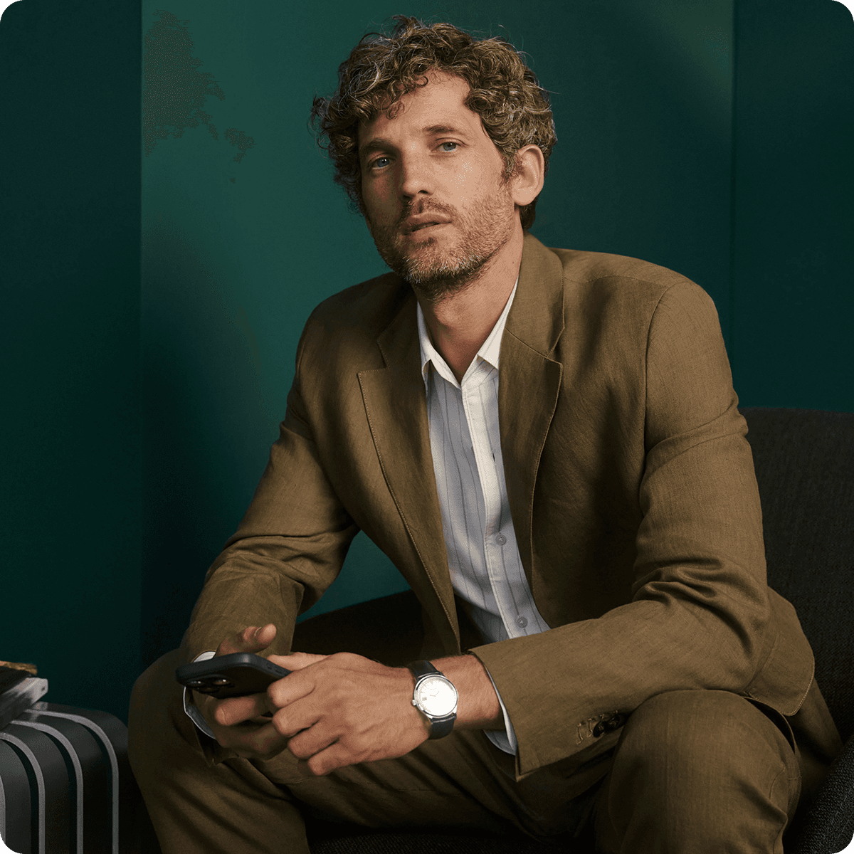 Man with curly hair in a brown suit sits on a chair, holding a smartphone, against a dark green background.