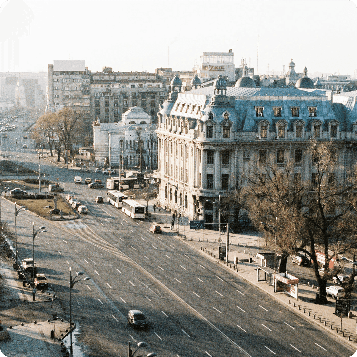 Aerial view of a wide city street with cars, buses, and historic buildings on a clear day. Trees and distant urban landscape are visible.