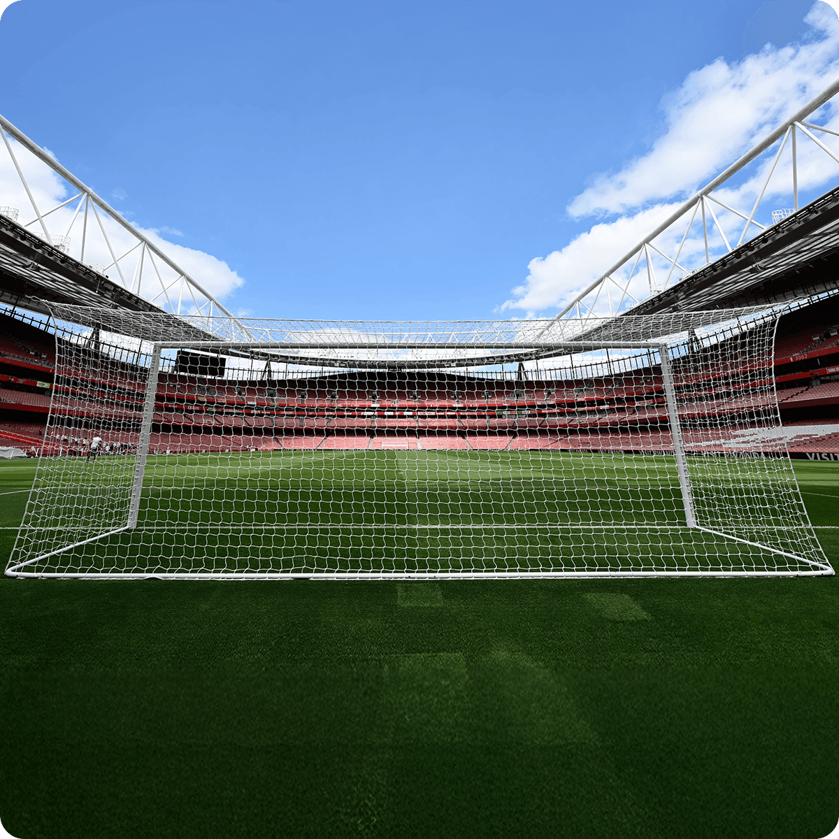 View of an empty soccer stadium with a goalpost in the foreground, green field, and blue sky with clouds above.