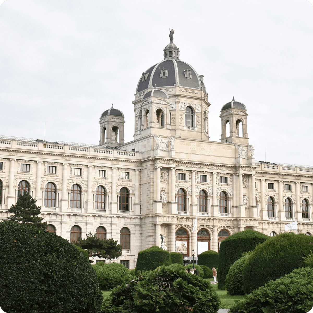Historic building with a grand dome and ornate facade, surrounded by manicured greenery and a cloudy sky in the background.