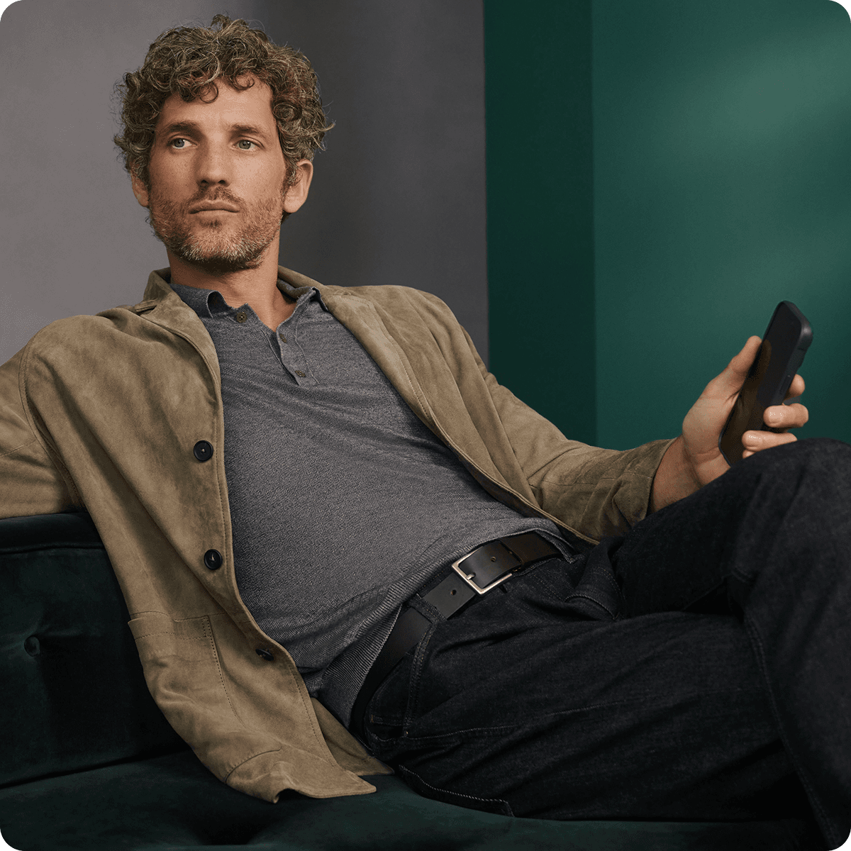 Man with curly hair in a beige jacket and gray shirt, holding a smartphone, reclines on a dark green sofa against a green and gray backdrop.