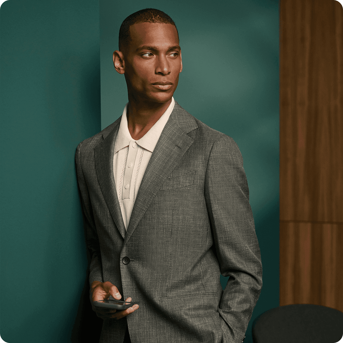 Man in a gray suit and white shirt holding a phone, standing against a green and wood-paneled background, looking to the side.