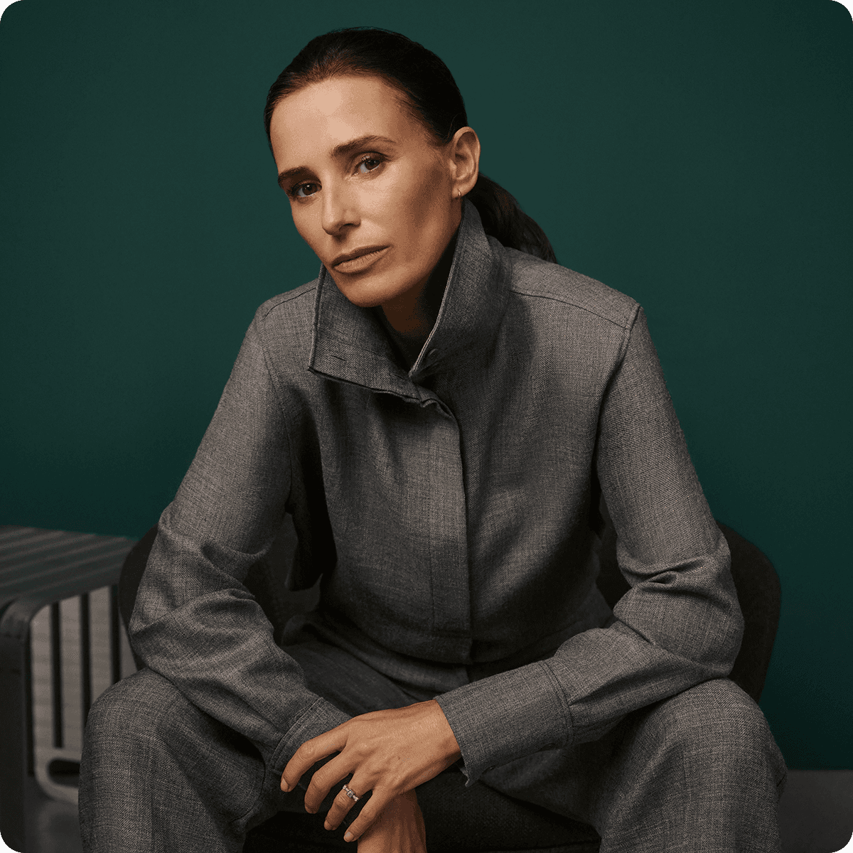 A person in a gray suit sits confidently against a dark green background, with a neutral expression and hands resting on their knees.