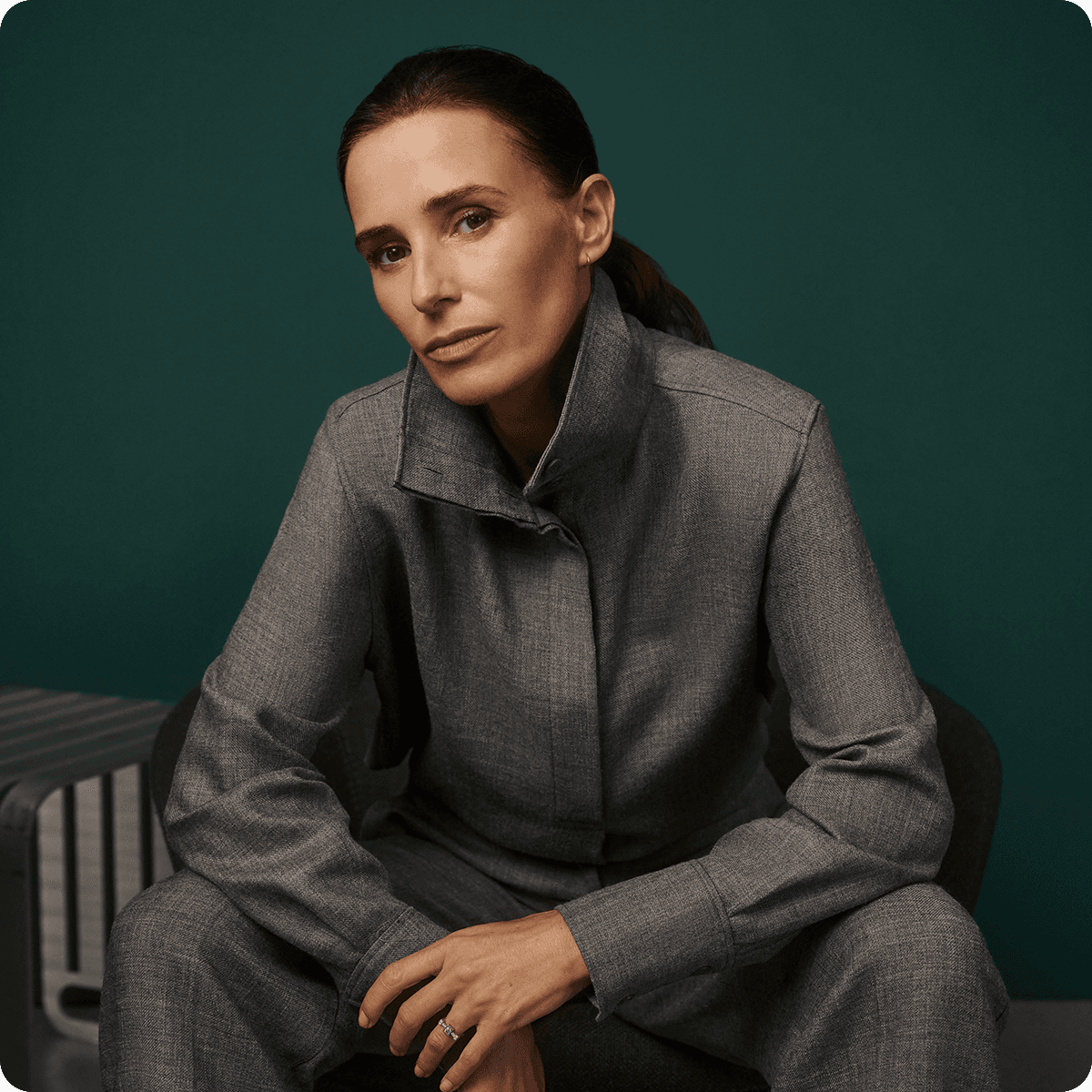 Woman in a gray suit sits confidently on a chair against a dark green background, with a serious expression and hands resting on her knees.