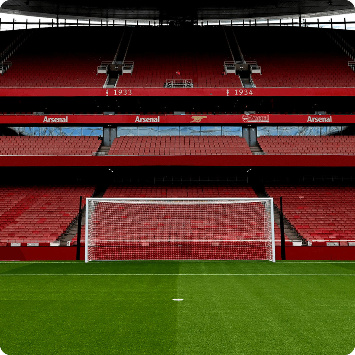 Empty football stadium with red seats, goalpost centered on green field, and banners displaying "Arsenal" in the background.