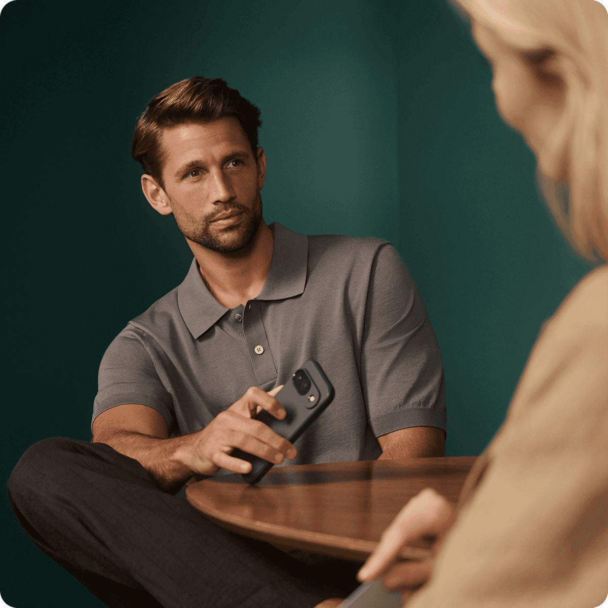 Man in a gray polo shirt holding a phone, sitting at a table, looking at a blurred person in the foreground.