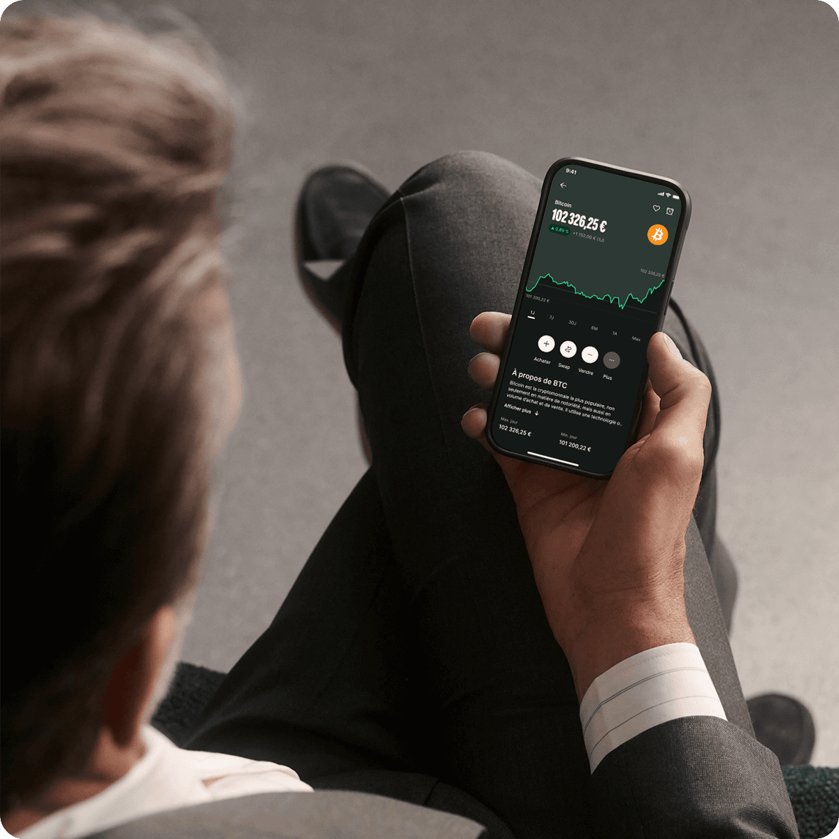 Person in a suit holding a smartphone displaying a cryptocurrency trading app with graphs and data.