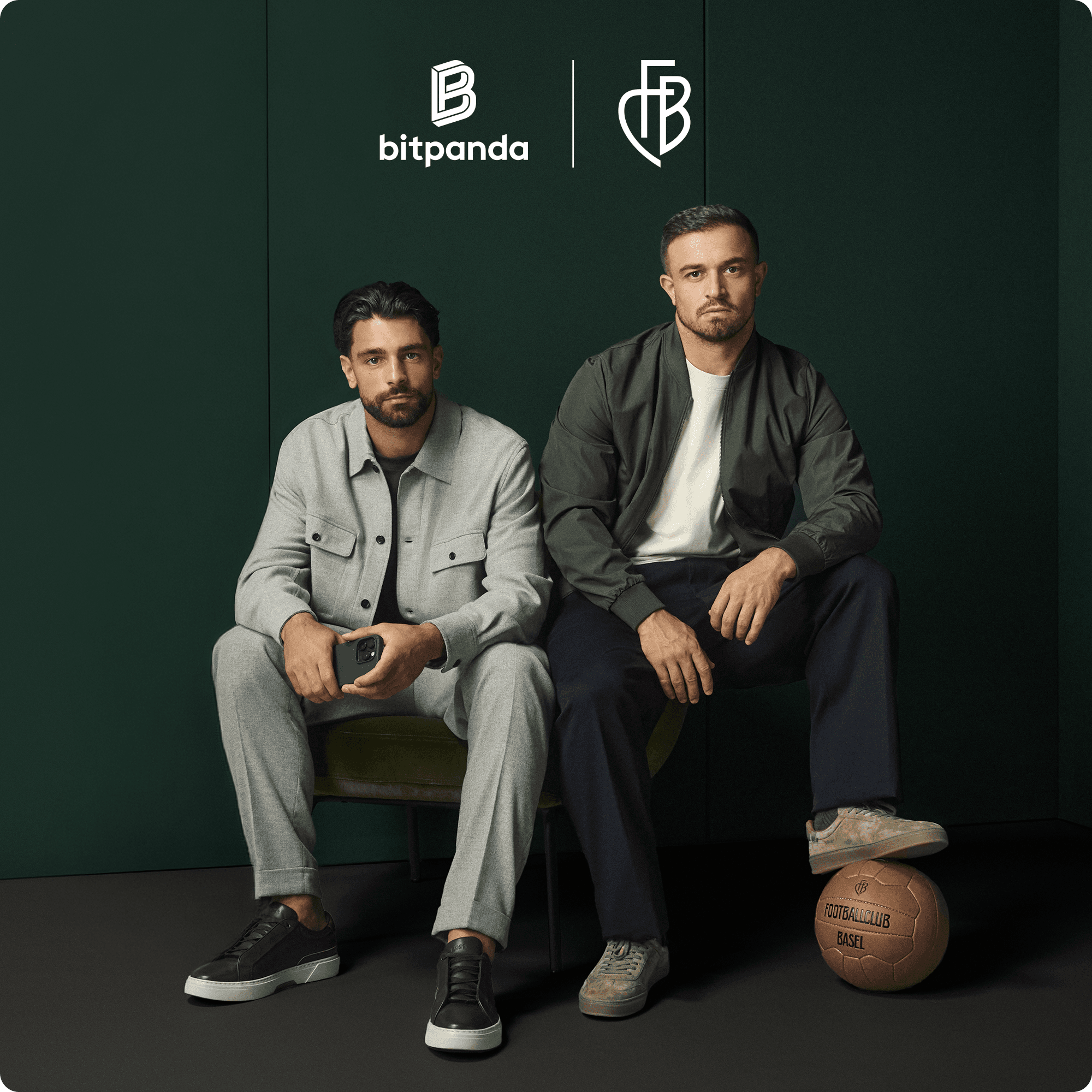 Two men pose seated in front of a dark green backdrop, one holding a phone, the other resting a foot on a vintage leather football.