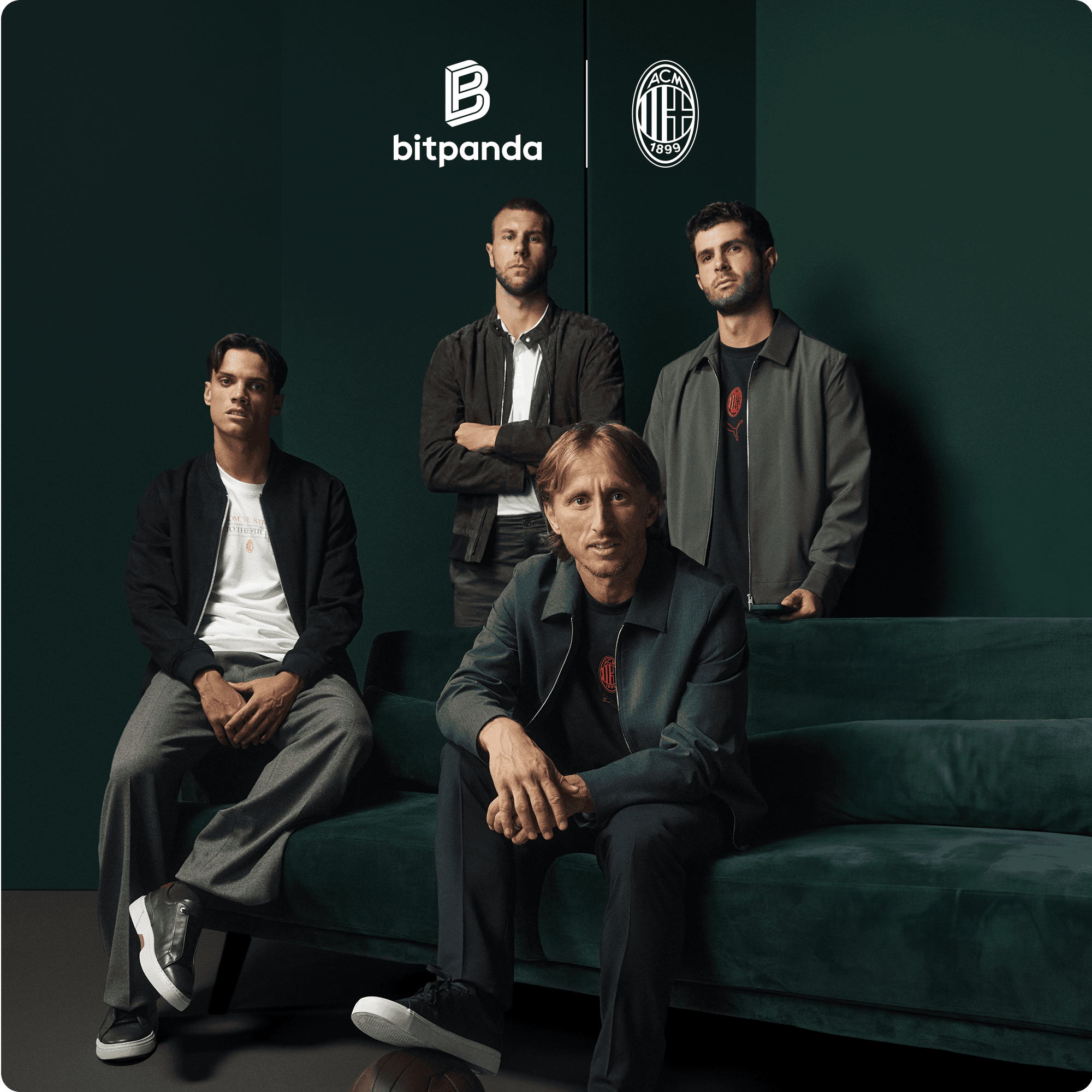 Four men in casual jackets pose around a dark green sofa with Bitpanda and AC Milan logos on the dark green wall behind them.