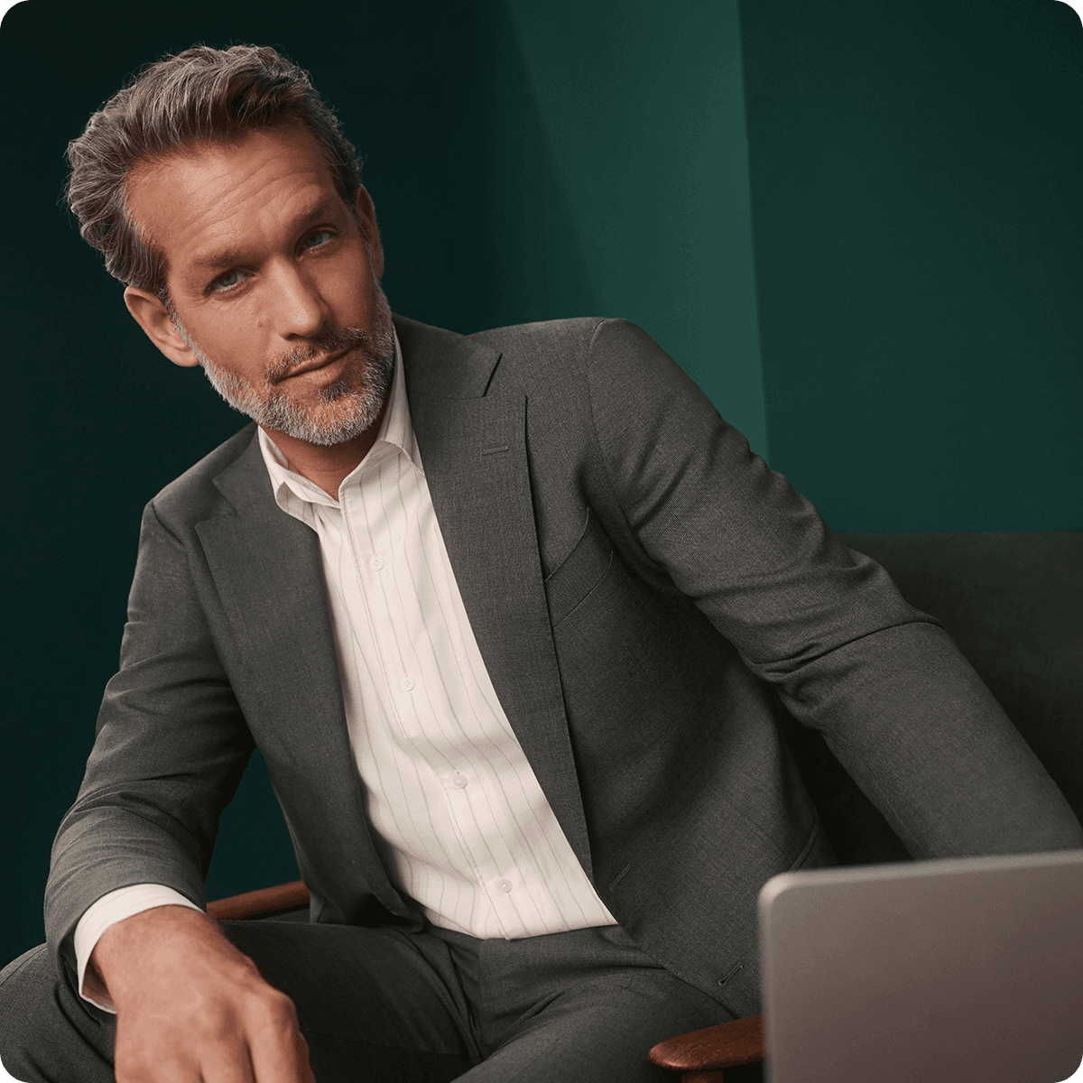 Man in a gray suit and white shirt seated, looking at a laptop, with a teal background.